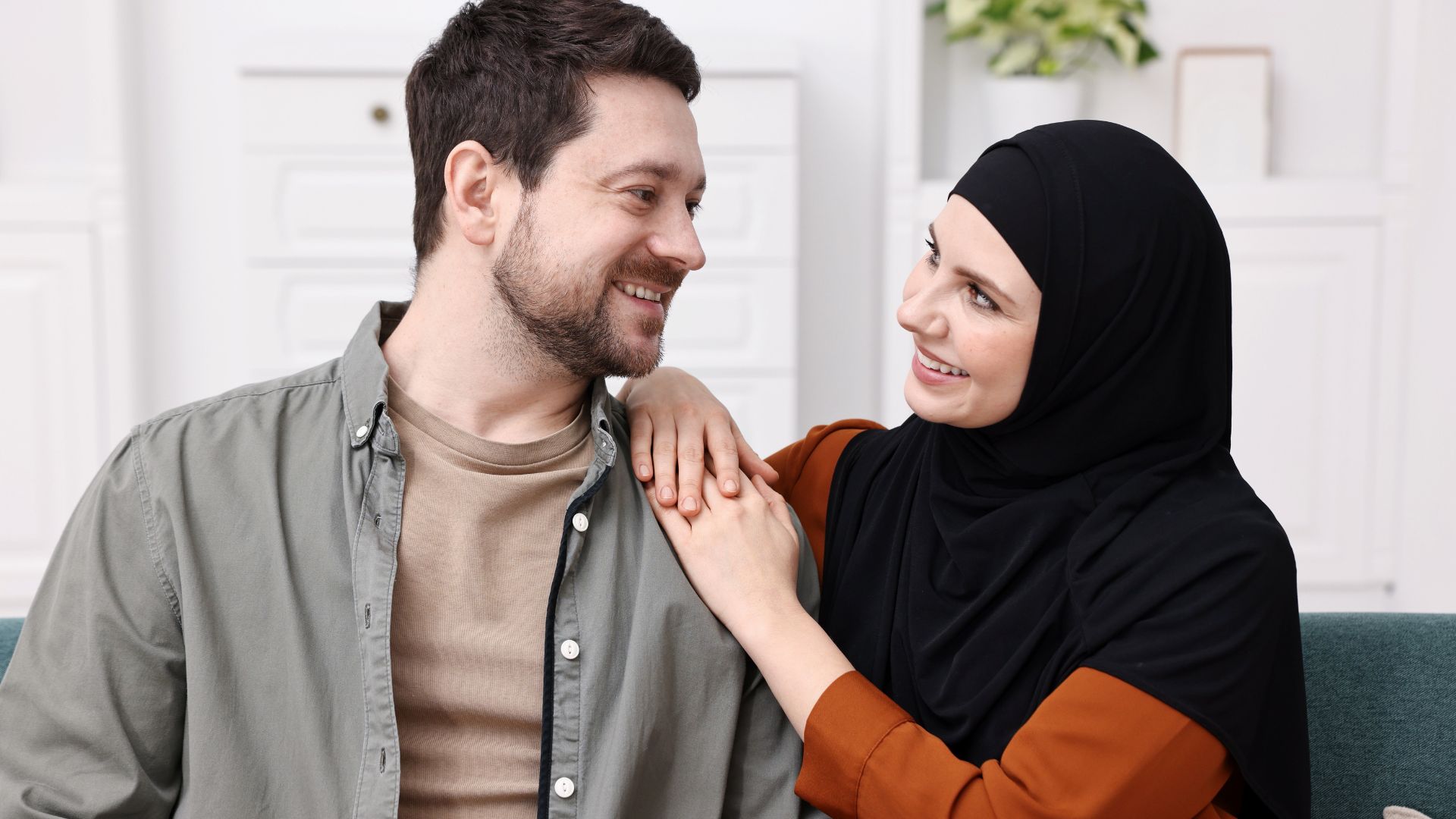 Smiling man and woman in a hijab share a warm moment; her hand rests on his shoulder.